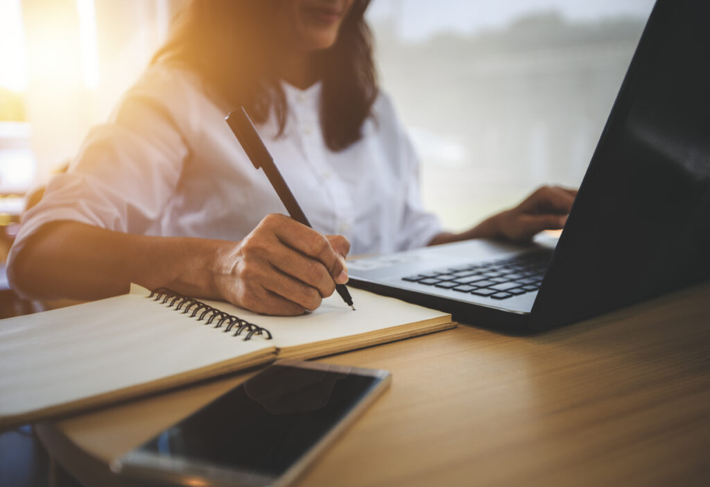 young woman with learning language during online courses using | Pegasus Online Woman focusing on her online business. Half of face is showing with hand on laptop, other hand holding pen, and writing on notebook with a phone in front.
